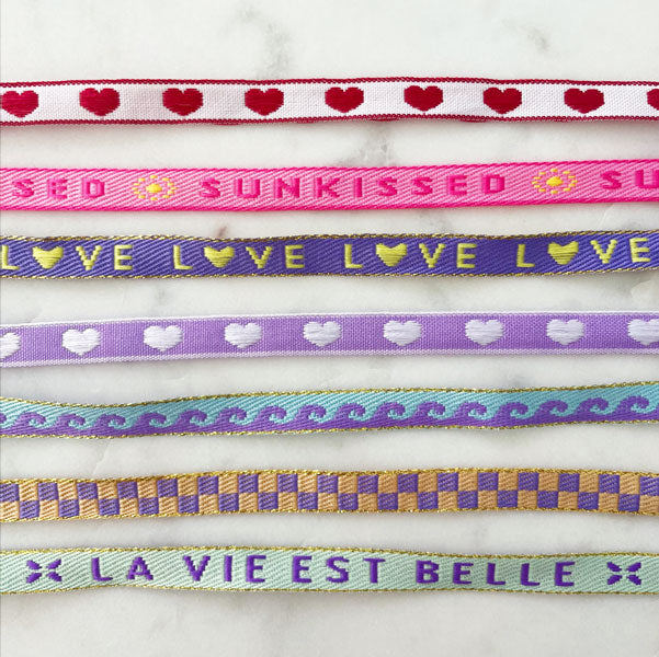 Gekleurde armbandjes met tekst om helemaal blij van te worden. Op het lint staat o.a. Sunkissed, Love, La vie est belle. 