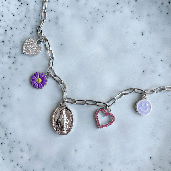 Een zilveren bedelketting met gekleurde bedels waaronder een lila smiley, bloem, hartje en een maria.